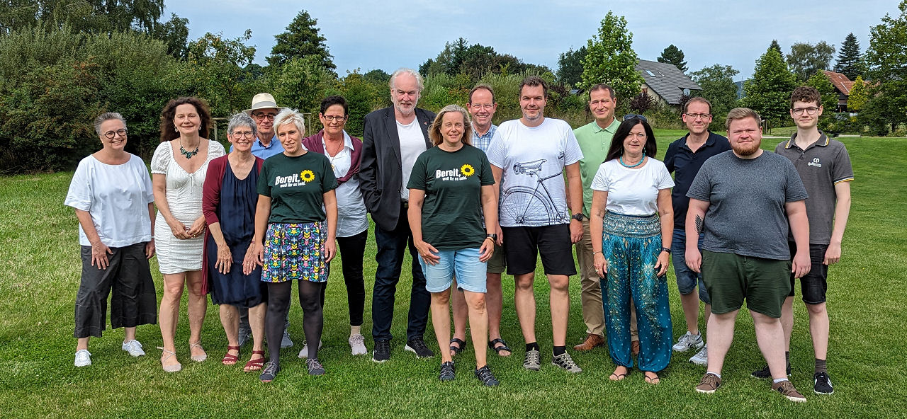 Ein Gruppenbild mit mehreren Menschen auf einer Rasenfläche vor eine Hecke aus Büschen und Bäumen.