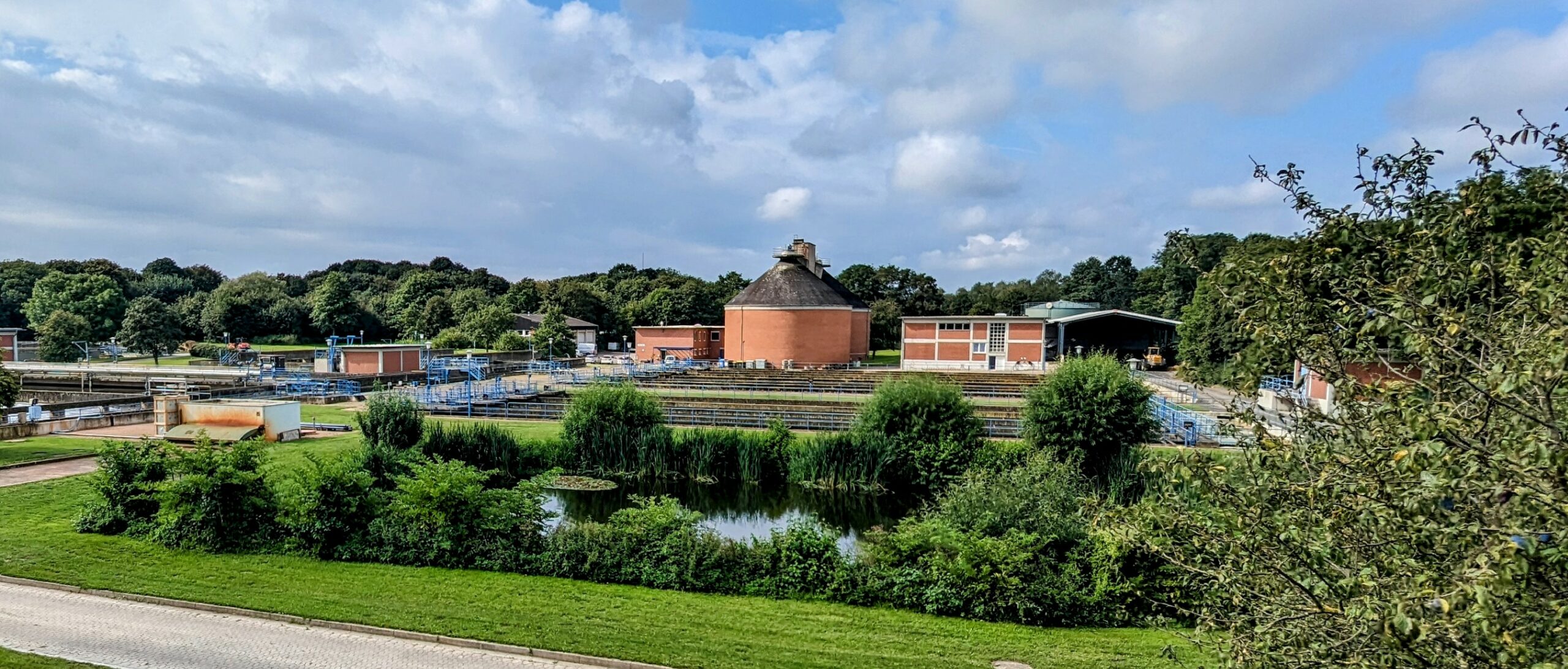 Im Vordergrund ein von Büschen und Bäumen ums#umter Teich, im Hintergrund sieht man einen Faulturm der Kläranlage und einige niedrigere Betriebsgebäude.