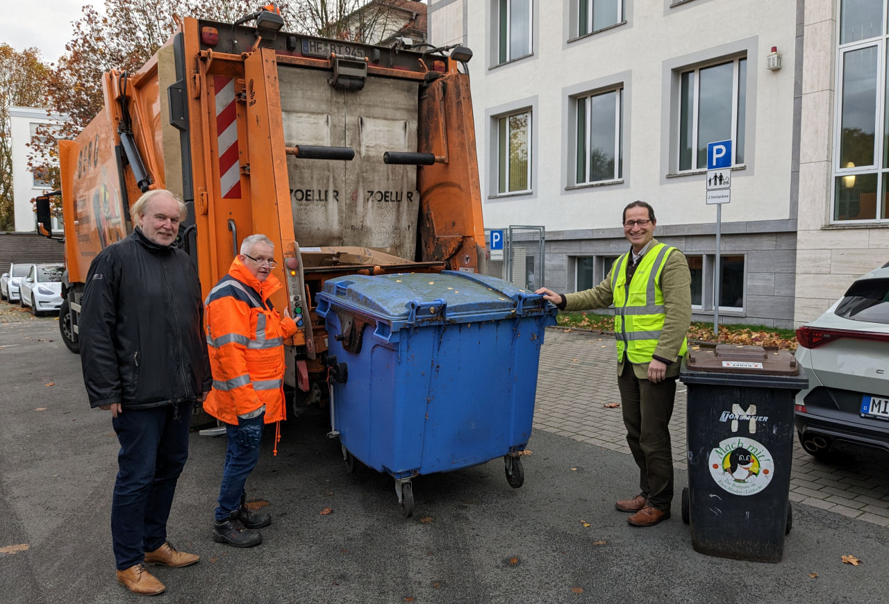 Ein Müllfahrzeug mit einer großen blauen Tonne am Heck. Drei Männer haben sich um das Fahrzeug gruppiert und schauen in die Kamera