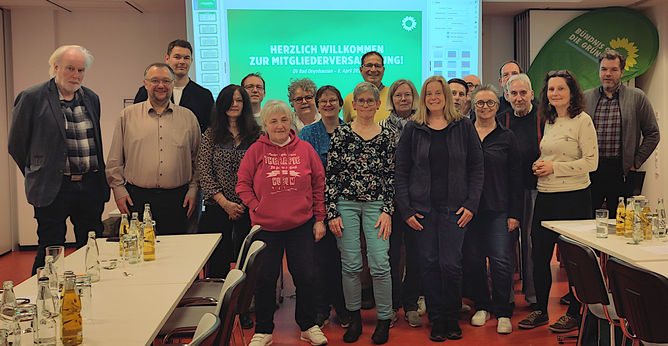 Ein Gruppenbild der Kandidatinnen und Kandidaten der GRÜNEN in Bad Oeynhausen. Rechts und links sieht man die Enden von Tischreihen mit einigen Getränken darauf, dahinter eine Gruppe von Frauen und Mämnnern vor einer zur Hälft von der Gruppe verdeckten Leinwand mit einer Präsentation der Grünen.
