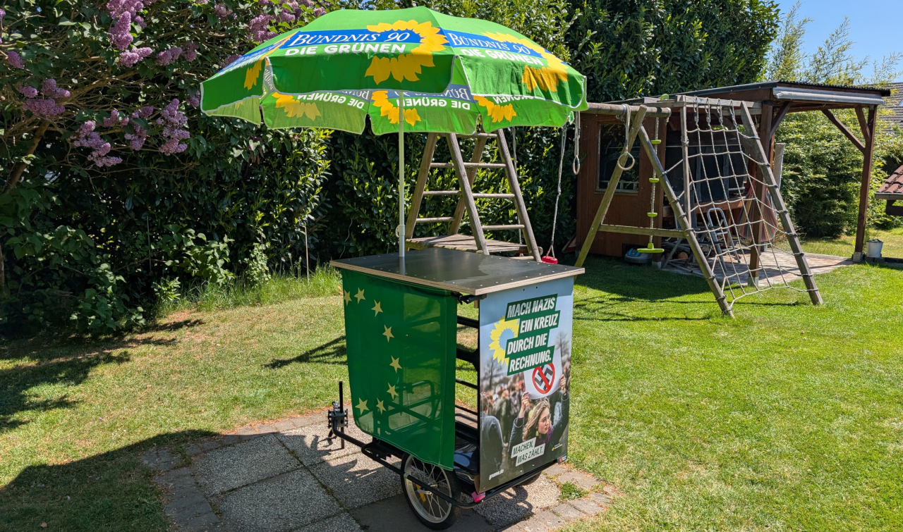 Ein Fahrradanhänger als Wahlinfostand mit einem Stehtisch und integriertem Sonnenschirm inkl. Werbung für die Grünen.