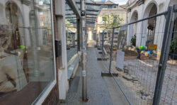 Eine Baustelle in einer Fußgängerzone. Die zur Verfügung stehende Verkehrsfläche ist mit einem hohen Bauzaun abgesperrt und sehr schmal. Links sieht man die Schaufenster einiger Geschäfte.