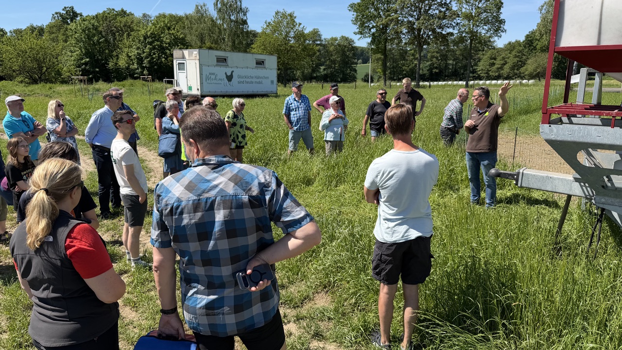 Eine Gruppe von Menschen steht auf einer grünen Wiese mit hohem Gras. Rechts sieht man die Deichsel eines großen Anhängers, im Hintergrund steht auf der Wiese ein mobiler Hühnerstall. Ein Mann deutet auf den zu der Deichsel gehörigen Wagen - welcher nicht im Bild zu sehen ist - und scheint etwas zu erklären.